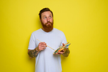 Undecided man with beard and tattoo is ready to draw with brushes