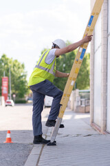 telecommunications technician looking down climbing a yellow ladder wearing a reflective vest, boots and hard hat 