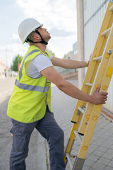 telecommunications technician holding a ladder looking upward, wearing a safety helmet and reflective vest