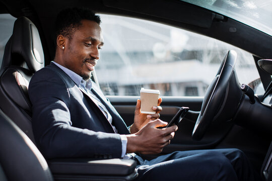 Handsome african man in business suit sitting on driver's seat of his luxury car, drinking coffee and using modern smartphone. Young male taking stop for checking work emails on mobile. - Powered by Adobe