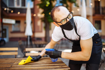 A waiter in a protective mask and a glove disinfects furniture in a street cafe. Prevention of coronavirus infection. Coronavirus prevention concept.