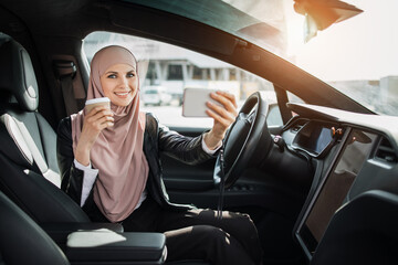 Positive business woman in hijab having video call on smartphone while sitting in luxury electric car. Attractive lady drinking fresh coffee and talking on mobile.