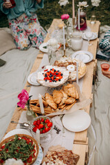 outdoor picnic feast. festive pallet table with desserts and snacks. people sitting near the table with food, drinks and  candles.