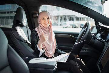 Pretty arabian woman in hijab smiling and showing thumb up while sitting in luxury electric car with modern laptop on knees. Concept of business, technology and transport.