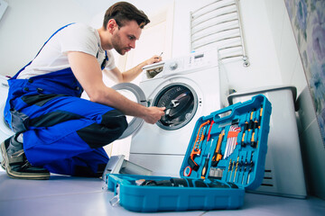 The young handsome repairman in worker suit with the professional tools box is fixing the washing machine in the bathroom - obrazy, fototapety, plakaty