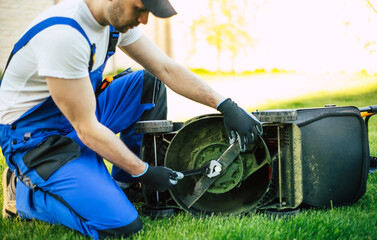 Workman in the suit is repairing lawn mower with help of a tool outdoors - obrazy, fototapety, plakaty