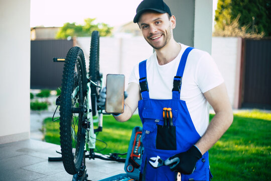 Handsome smiling bicycle repairman shows smartphone on the camera on the mountain bicycle background