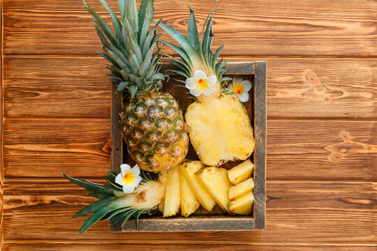 Sliced Pineapple. Tropical summer fruit pineapple halves and whole pineapple on brown dark table in wooden box with tropical plumeria flowers. Top view