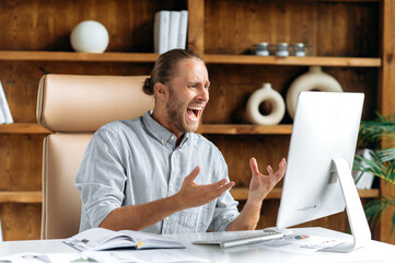 Frustrated angry caucasian businessman or CEO with a beard sits in the office, using a computer,...