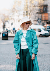 woman in sunglasses, hat and turquoise trench walking in English city 