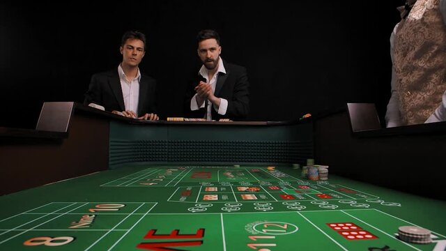 Two men in business suits stand at a green craps table in a casino. The player rolls the dice and wins. Males rejoice with delight and wow emotions. View from inside of game table. Slow motion.