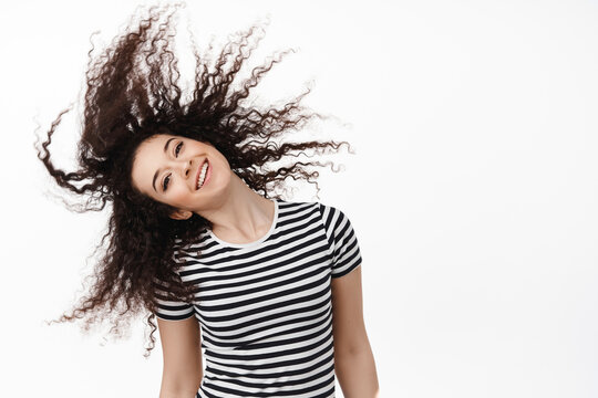 Portrait Of Happy Natural Brunette Woman Flip Curly Hair And Smiling Carefree, Looking Cheerful, Standing Against White Background And Having Fun