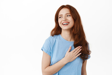 Happy candid redhead girl laughing and smiling naturally, holding hand on heart and looking aside...