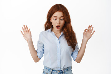 Dropped something. Shocked redhead girl looking down at surprising thing, checking out advertisement below, standing in blouse against white background - 437996145