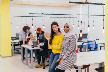 Portrait of two African American businesswomen talking to each other while standing in a modern business office with their colleagues, coworkers in the background. Multi-ethnic society..
