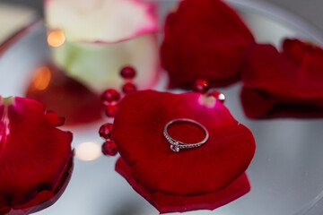 Wedding ring lies on red rose petals on the background of candles. Marriage proposal. Romantic atmosphere.