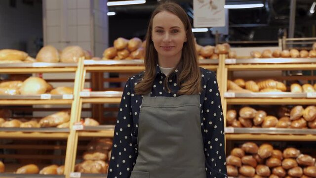 Woman who owns a small bakery in an apron smiles in her candy store. A woman looks at the camera on the background of bread
