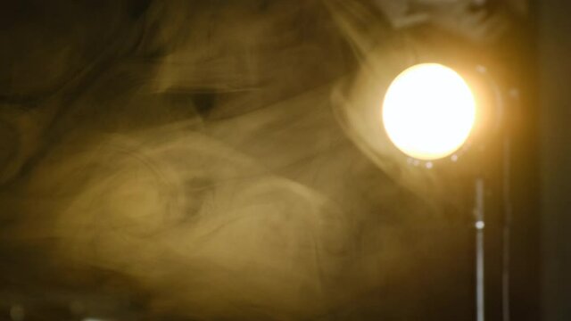 Video light of yellow light 2700 Kelvin stands on rack with fresnel lens attachment and shines on smoke that is around lamp. Focus on smoke, great background footage.