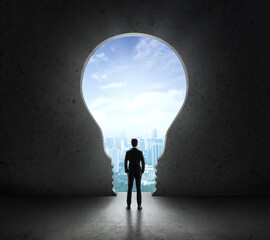 Fototapeta na wymiar Businessman standing in front of bright big light bulb shining door in a concrete wall with a view to the city