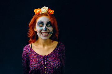 A young woman in day of the dead mask skull face art. Woman with skull makeup and red hair showing teeth on dark background with copy space