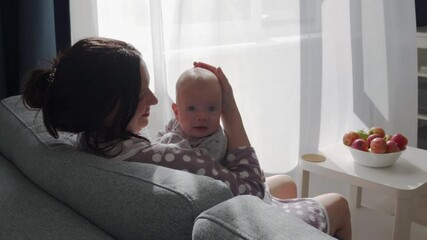 Mother holding hugging kissing her baby sitting on the sofa in the living room on a sunny day. High quality 4k footage