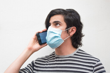 Portrait of a young man with medical face mask talking on the phone and looking up, whit background and striped t-shirt. Scene of COVID or coronavirus pandemic lifestyle.