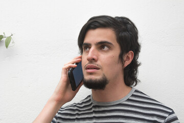 Portrait of a young man with beard doing a call or talking on the phone and looking up, white background and striped t-shirt. Communications on smartphone scene