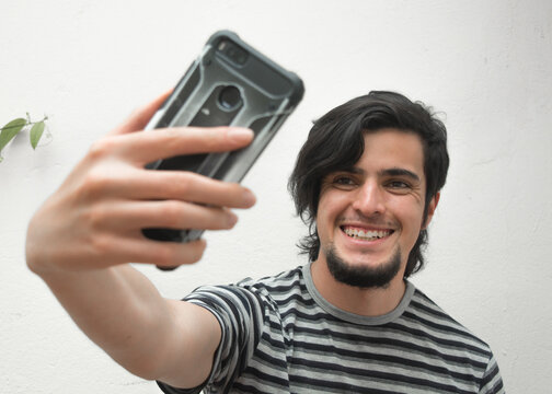 Portrait of a young man with beard holding a phone on his hand smiling and taking a selfie. Model wearing a black and white striped t-shirt. Happiness and confidence face or facial expressions.