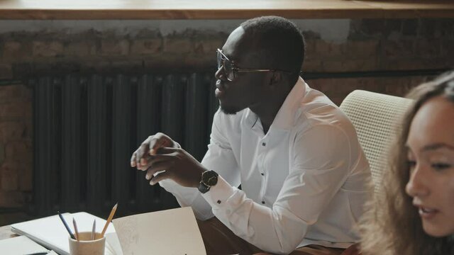 Side-view medium slowmo of young handsome African American businessman in white shirt and eyeglasses discussing new project with colleagues sitting at table in modern loft coworking office