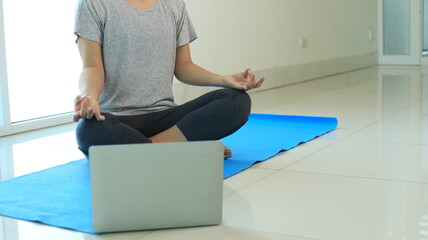 Woman workout yoga through laptop. She sits on blue yoga mat. Female is meditating for ready to exercise yoga.