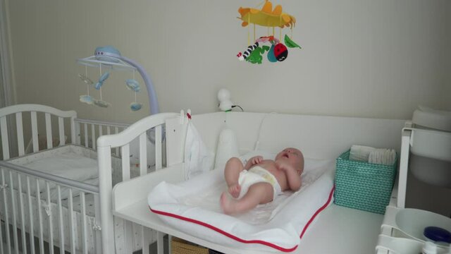 Baby in a diaper on white changing table looks at the toy mobile above him, 3 month old baby boy. High quality 4k footage