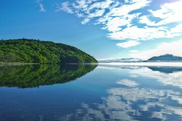 晴れた日の雲の浮かぶ爽快な空と湖畔の森を反射する静水の湖。