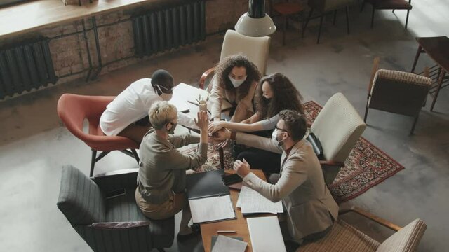 Top-view slowmo of young diverse team of coworkers wearing protective masks sitting at work table in modern loft workspace then putting hands together in middle Concept of teamwork and partnership