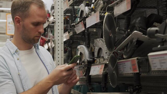 Man chooses a circular saw using a smartphone in a tool shop. Male chooses a product in a hardware store.