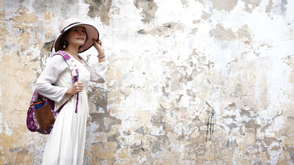 Woman with white hat on grunge wall as tourist in white dress.