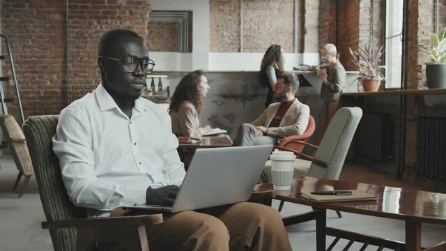 Medium slowmo of concentrated young African-American man in stylish white shirt and eyeglasses working on laptop in cozy loft-style coworking space while his colleagues chatting in background