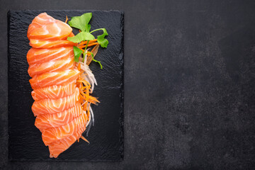 top view of fresh salmon sashimi fillet with carrot, radish slice and celery in black slate on dark grey texture background with copy space for text