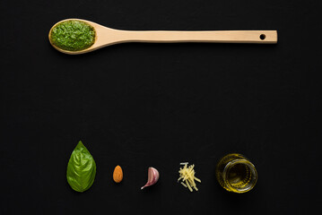 Top down view of basil pesto sauce in a wooden spoon and a basil leaf, almond, garlic, parmesan cheese and olive oil as ingredients on a black surface with copy space