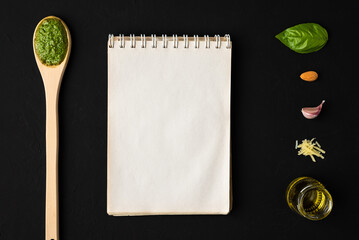 Basil pesto sauce on a wooden spoon, with a basil leaf, almond, garlic, parmesan cheese and olive oil as ingredients, and a notebook with blank paper for text