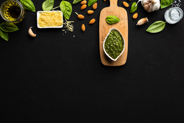 Pesto sauce in a white leaf-shaped bowl and a little wooden table with basil leaves, parmesan cheese, almonds, garlic, olive oil and salt as ingredients on a black surface with copy space