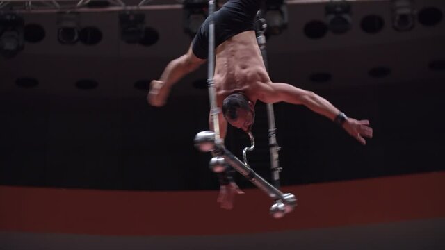 Acrobatic man training at the circus arena - hanging upside down on the rotating performance construction holding on it with his mouth
