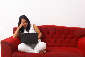 Obraz premium Asian woman using a laptop while sitting on red sofa.Asian woman using a laptop while sitting on red sofa. Work from home concept