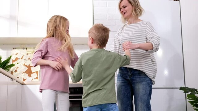 Excited mom or babysiter and two kids boy and girl jumping, dancing laughing in modern scandinavian house kitchen. Happy family mother daughter son having fun enjoy playing singing together at home.