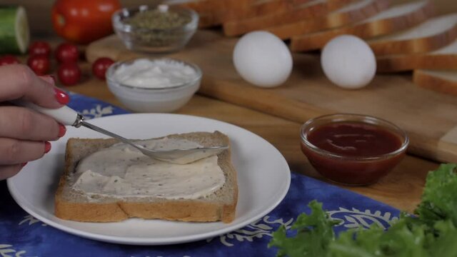 Female hands spreading a spoonful of mayonnaise on a slice of brown bread -making sandwich . Closeup shot of two eggs  tomato ketchup  lettuce leaves  and white bread slices kept together on a wood...