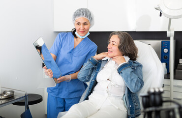 Satisfied elderly woman patient looks in the mirror after the procedure, sitting on the couch in the cosmetology office