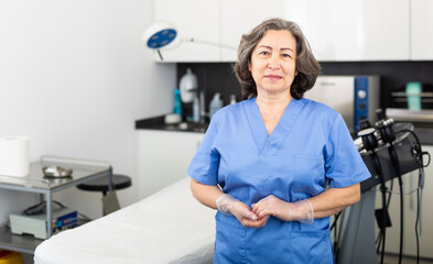 Portrait of confident professional elderly woman cosmetologist wearing blue uniform standing in modern medical aesthetic office