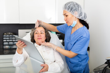 Asian female cosmetologist giving professional advice to elderly woman before hardware facial skin treatments in cosmetological office .