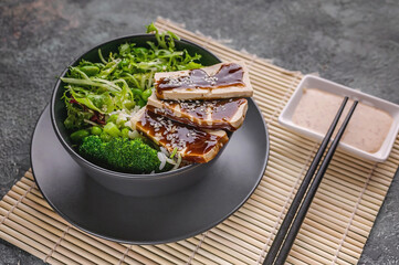 Pork meat with a mix of fresh tasty and healthy vegetables, boiled rice, teriyaki sauce in a bowl. Gray crockery on a bamboo mat, black chopsticks, peanut sauce. Still life of traditional asian food