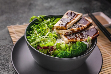 Close-up pork meat with a mix of fresh tasty and healthy vegetables, boiled rice, teriyaki sauce in a bowl. Gray crockery on a bamboo mat, black chopsticks, peanut sauce. Traditional asian food