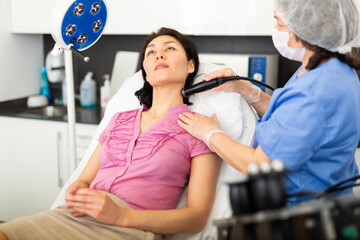 Obraz premium Highly qualified cosmetology doctor makes a young female client sitting in a couch vacuum cleaning of the face with ..the help of a device in the clinic room